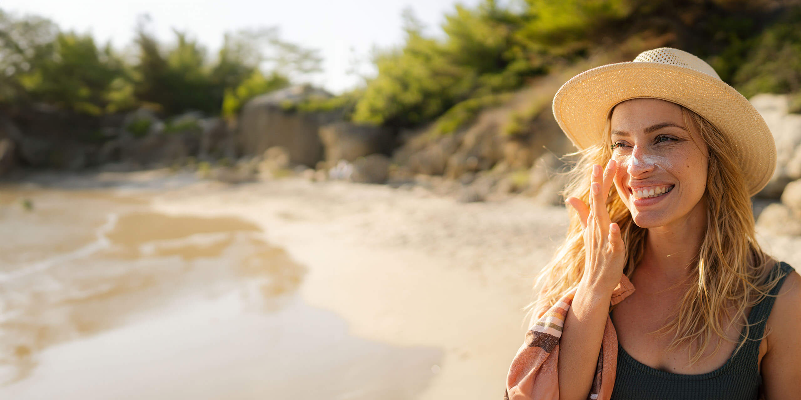 Eine Frau an einem sonnigen Strand trägt Sonnencreme auf ihre Nase auf. Sie trägt einen Strohhut, ein dunkles Tanktop und hat ein Handtuch über der Schulter. Im Hintergrund sind Sand, Felsen und Grün zu sehen. Sie lächelt und schaut zur Seite.