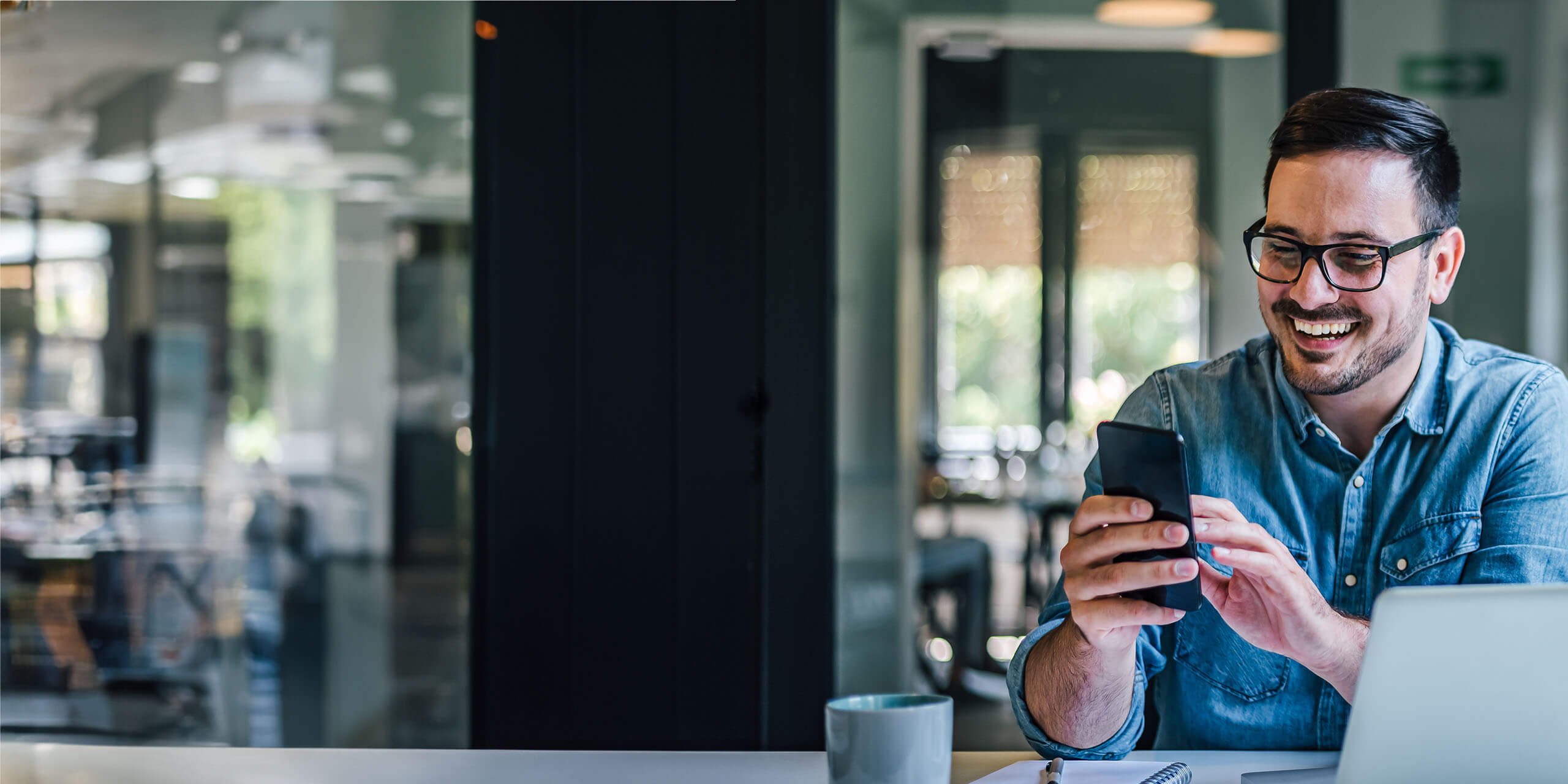 Ein fröhlicher Mann mit Brille und Jeanshemd sitzt an einem Schreibtisch in einem hellen Büro, benutzt sein Smartphone und lacht, neben ihm stehen ein Laptop und eine Kaffeetasse.