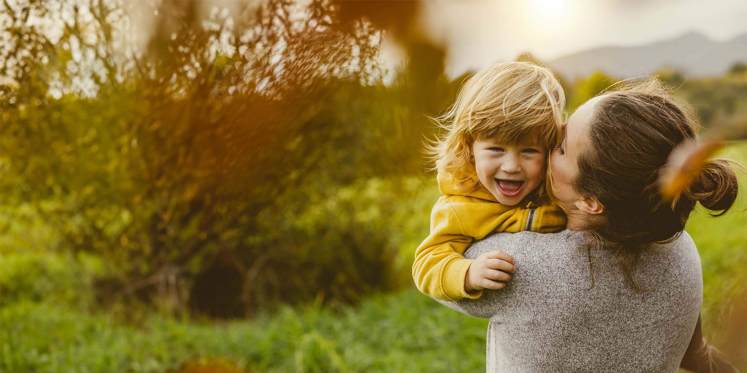 Ein fröhliches kleines Kind in einem gelben Hemd lacht, während es von seiner lächelnden Mutter im grauen Pullover gehalten und auf die Wange geküsst wird, draußen mit viel Grün und sanftem Sonnenlicht im Hintergrund.